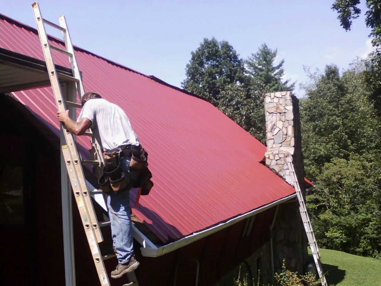 Roofer on a ladder beside a red metal roof and stone chimney — Soffit & Fascia Repair by Roofing Brothers in Pequannock